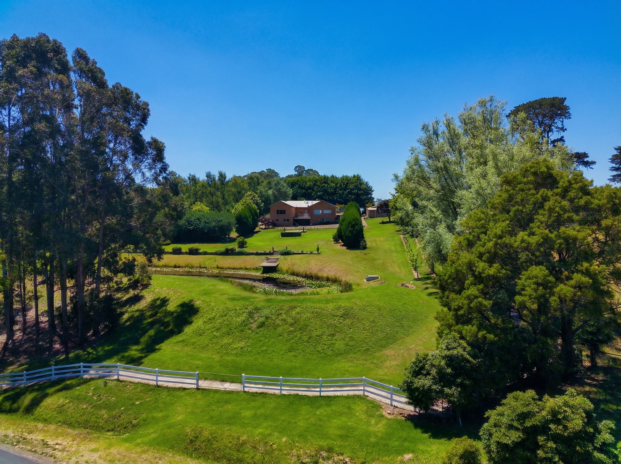 Aerial view of Springbank property from O'Malleys Road showing white post-and-rail fencing, sweeping manicured lawns, dam with merbau deck, mature eucalypts and willows, and the Mediterranean-style residence nestled among established gardens